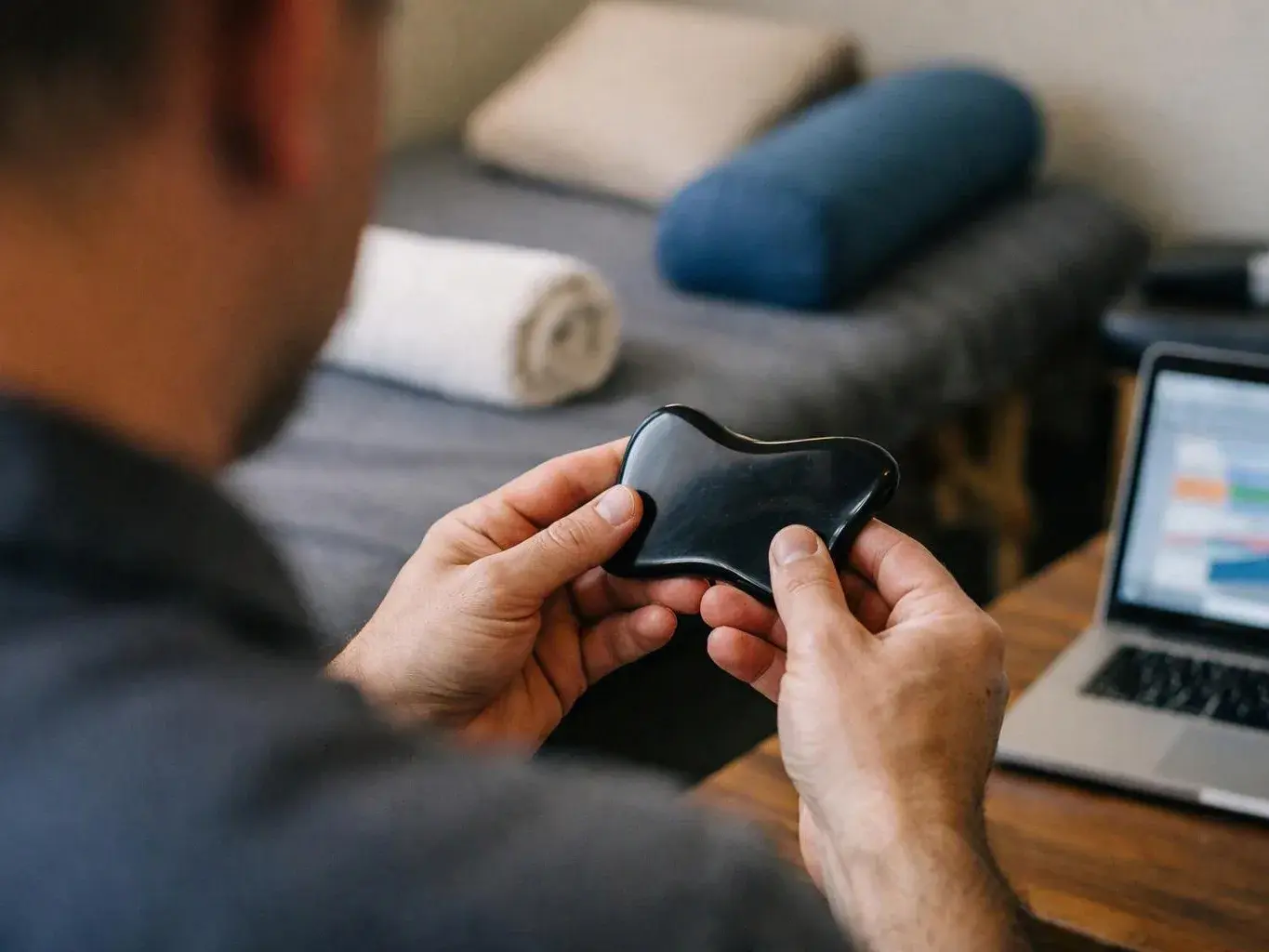 A person holds a small black massage tool beside a laptop in a bedroom or therapy room