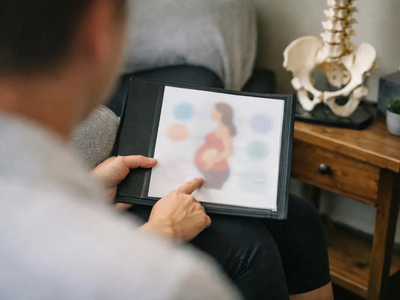 A person points at a blurred pregnancy illustration in a folder, with a spine model and side table behind them