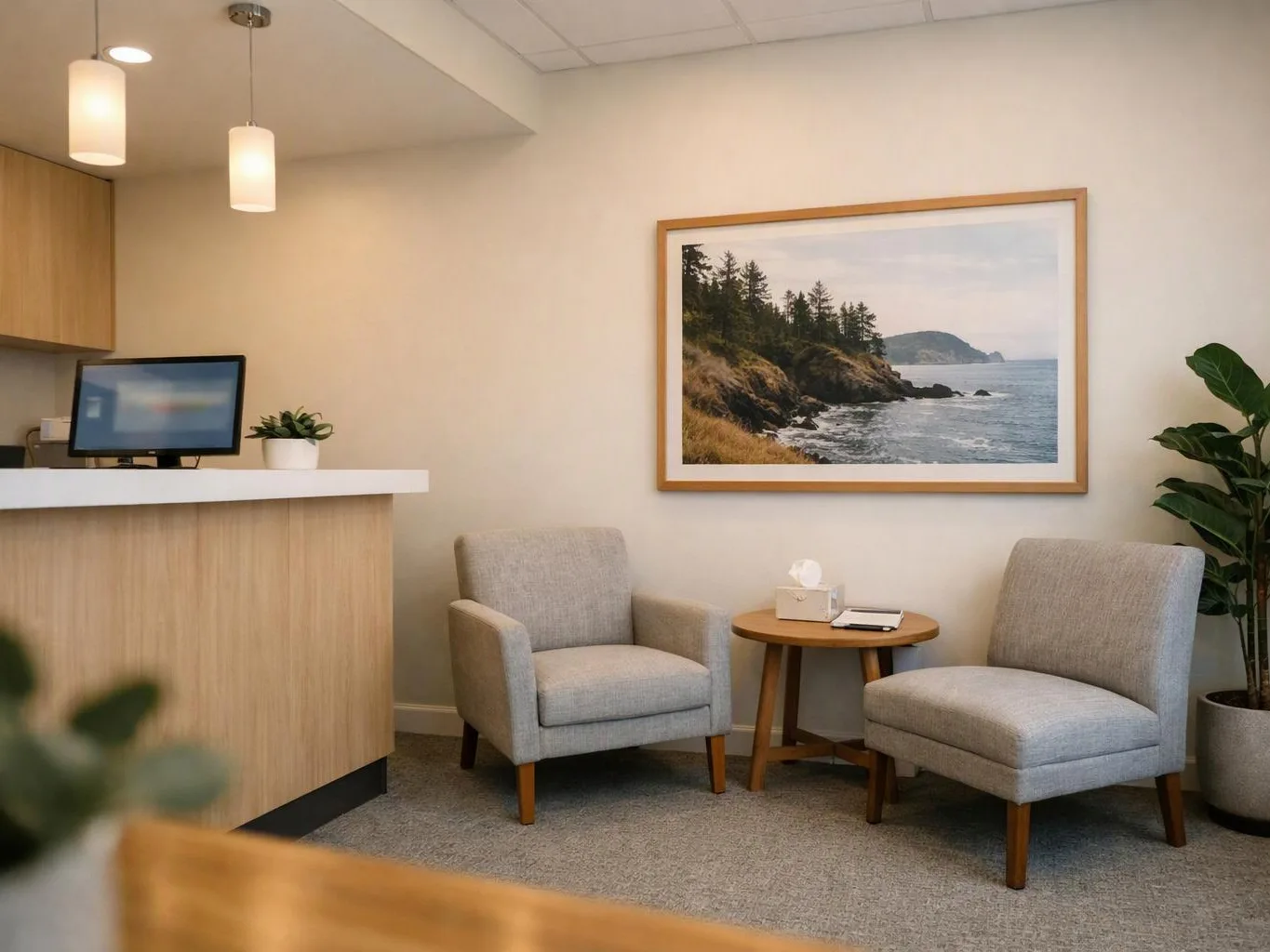 A quiet waiting room with two gray chairs, a small table, a reception counter, and a framed seascape on the wall